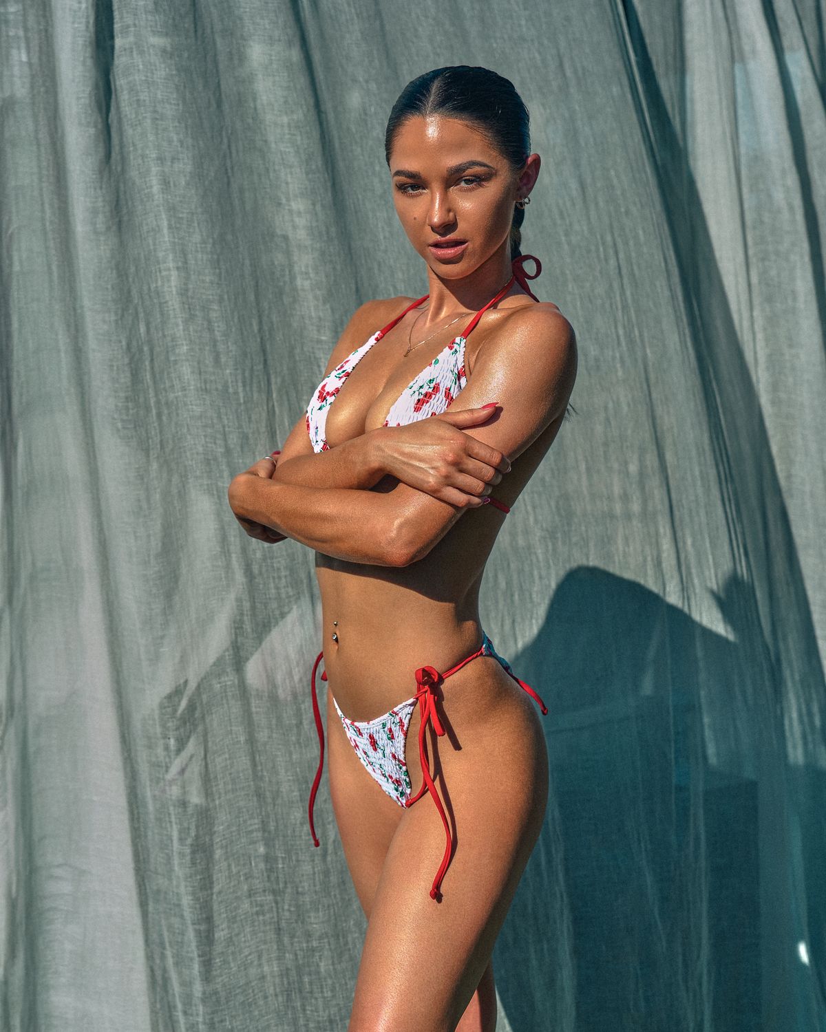 Model Lauren in a cherry-print bikini standing in profile against a sheer cabana curtain, arms crossed, sunlit, Dallas