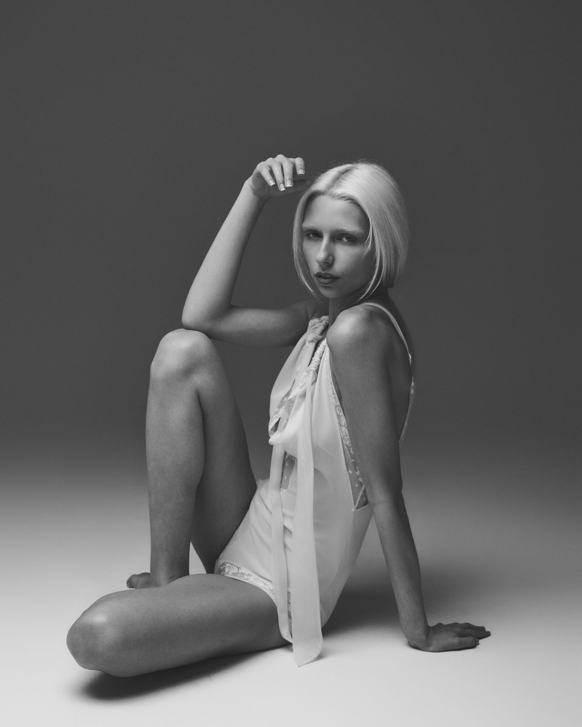Model Kalie seated on the floor with a knee raised, hand to her hair, sheer ivory lace bodysuit, black-and-white frame, Dallas