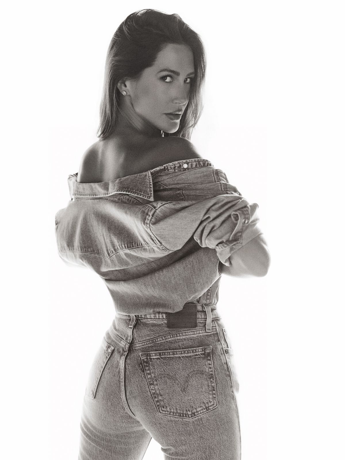 Giovanna from behind in dark jeans, denim eased off her shoulders, looking back at the camera in black and white.