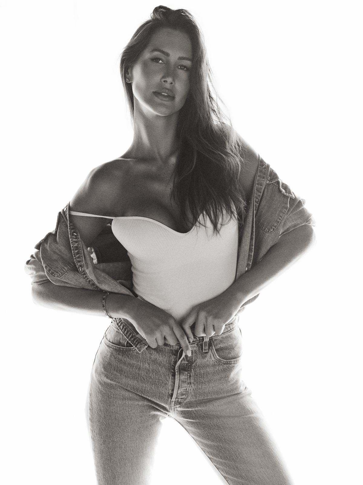 Giovanna standing in a white bodysuit with her denim jacket pushed to the waist, backlit black-and-white frame.