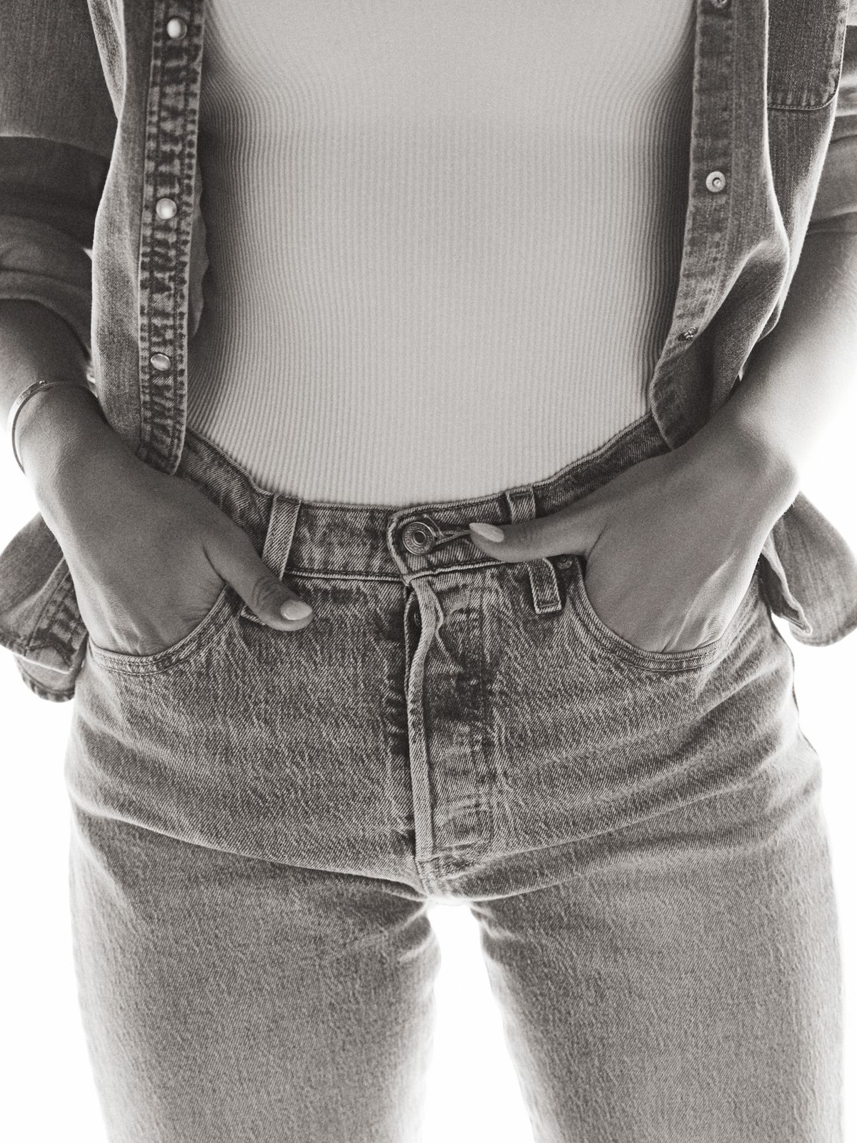 Black-and-white waist crop of Giovanna, hands tucked in her jean pockets under an open denim shirt.