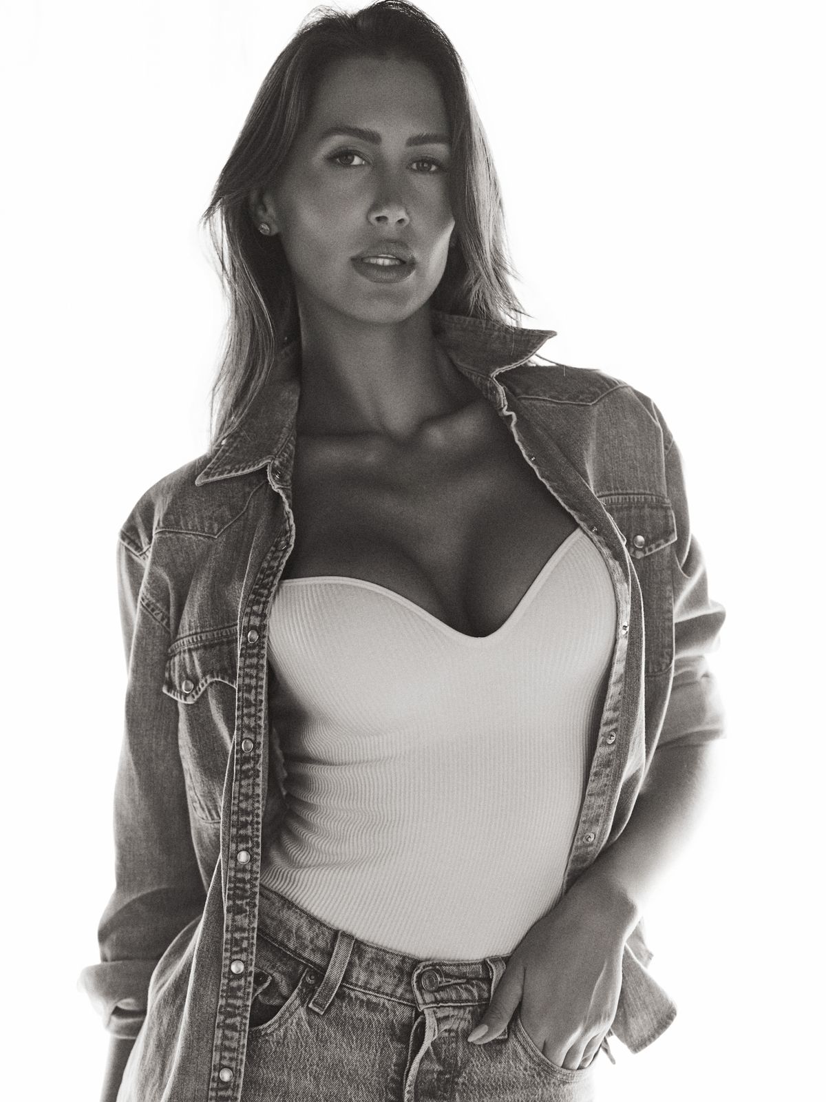 Backlit black-and-white portrait of Giovanna in an open denim shirt, chin lowered, hands in her jean pockets.